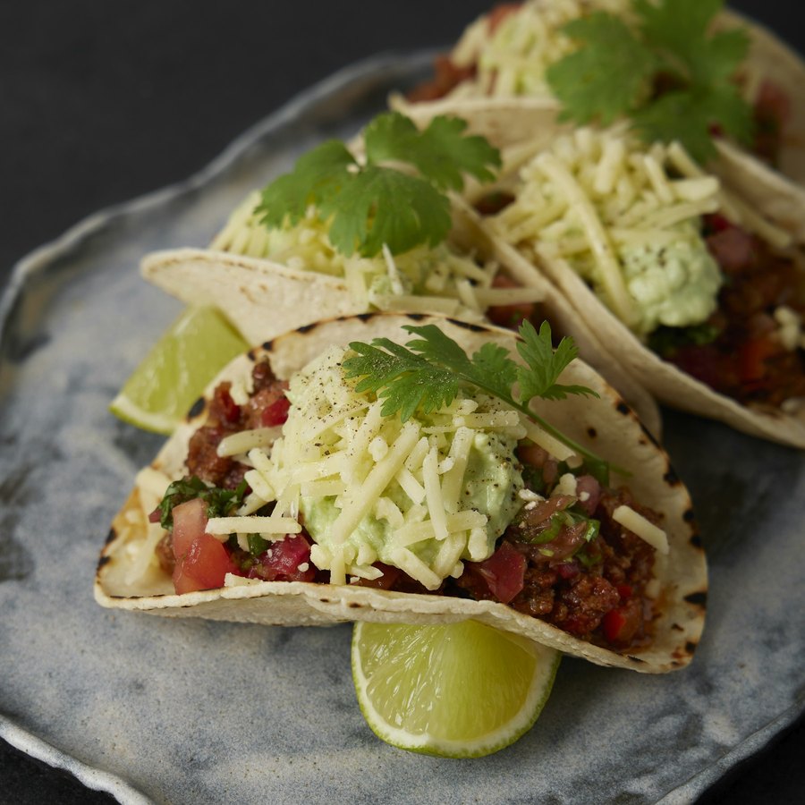 Three beef tacos with salsa, avocado crema, grated cheese, cilantro, and lime.