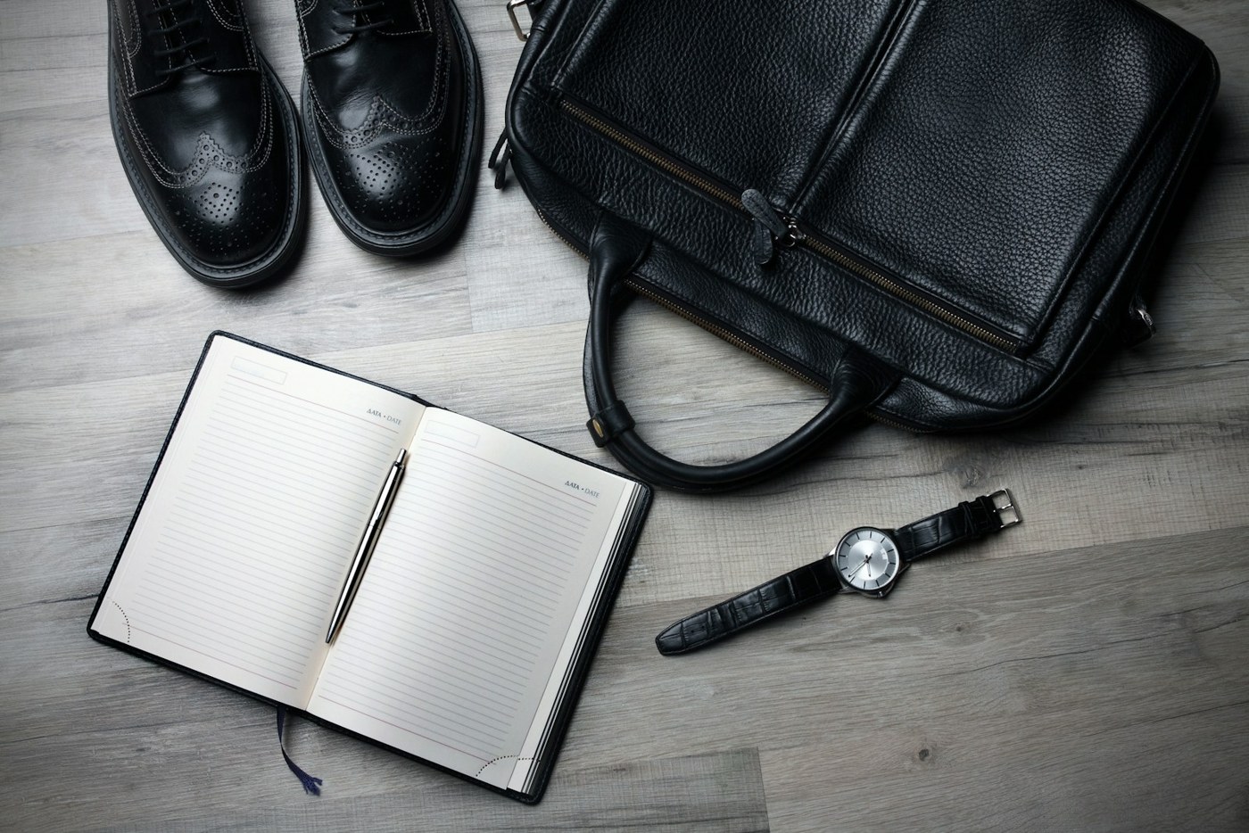 Everyday accessories, a notebook, shoes, and a leather bag on a pale wooden surface